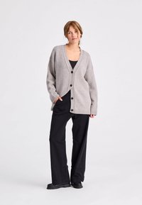 Grijze gebreide cardigan met een V-hals, knopen aan de voorkant en geribde manchetten, gecombineerd met zwarte broek met een brede pijp en forse zwarte schoenen.