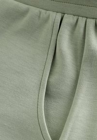 Zbliżenie na kieszeń jasnozielonego ubrania z widocznym szwem i miękką fakturą tkaniny pod elastycznym paskiem w talii.