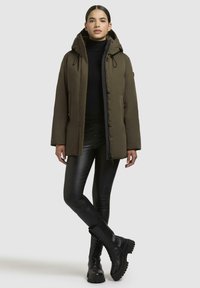 Olivgrüner gepolsterter Parka mit Kapuze, Reißverschluss und Knopfverschlüssen; getragen über einem schwarzen Rollkragenpullover und kombiniert mit schwarzen Lederhosen und Stiefeln.