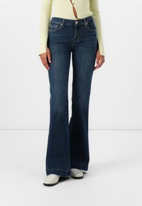 High-waisted flared jeans in dark blue denim, featuring classic pockets and a slight faded texture, paired with beige heeled boots.
