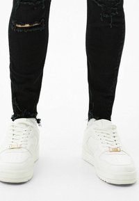 Svarta slitna jeans med fransade fållar, tillsammans med vita sneakers med en kraftig sula och guldaccent på snörningen.