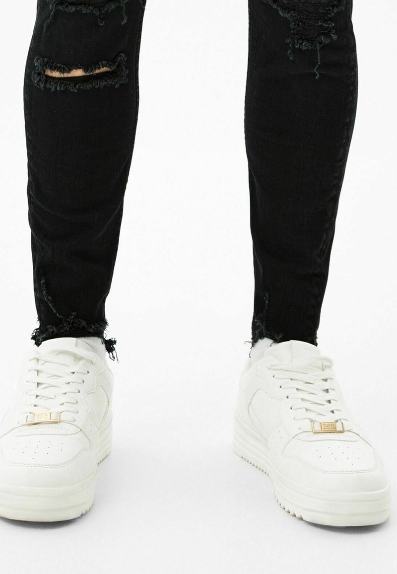 Svarta slitna jeans med fransade fållar, tillsammans med vita sneakers med en kraftig sula och guldaccent på snörningen.