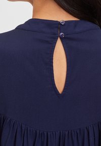 Retro di un vestito blu navy con due piccoli bottoni che chiudono un'apertura a chiave, mostrando parte della pelle e dei capelli scuri di una donna.