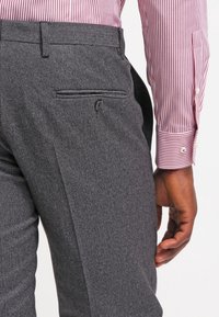 Graue maßgeschneiderte Hose mit strukturiertem Stoff, einer einzelnen Gesäßtasche und flacher Vorderseite, kombiniert mit einem rot-weiß gestreiften Hemd.