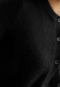 Detailní záběr na černý vlněný cardigan s knoflíky, který je nošen na horní části trupu, s viditelným výstřihem a třemi zapnutými knoflíky.
