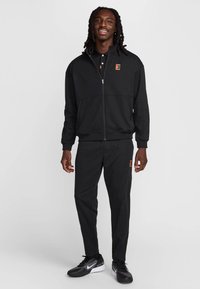 Μαύρο αθλητικό μπουφάν με φερμουάρ, πλαϊνές τσέπες και κεντημένο λογότυπο, σετ με μαύρο παντελόνι και μαύρα αθλητικά παπούτσια.