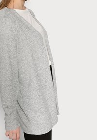 Cárdigan gris claro hecho de tejido texturizado, con un frente abierto, mangas largas y un ajuste relajado, llevado sobre una camisa blanca.