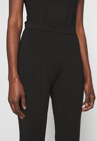 Närbild av en person som bär högmidjade svarta tajta byxor med synliga sömmar, samt en svart ärmlös topp, med avslappnade händer vid sidorna.