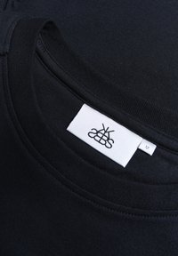 Schwarzes Baumwoll-T-Shirt mit Rundhalsausschnitt, das ein weißes Stoffetikett mit schwarzem Logo und der Größe 'M' auf der Innenseite aufgenäht hat.