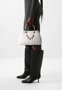 Biała pikowana torebka z złotym łańcuszkowym uchwytem, z czarnymi akcentami. Modelka ma na sobie szarą prążkowaną sukienkę i czarne kozaki za kolano.