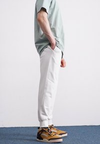 Tricou din bumbac de culoare verde deschis, pantaloni de trening albi cu buzunare și adidași maro cu accente negre, stând pe un fundal simplu.