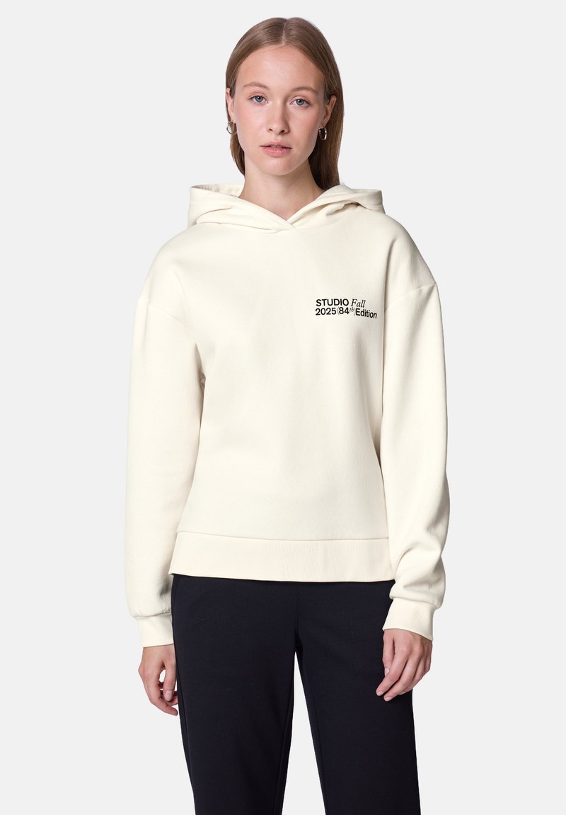 Felpa color crema in tessuto morbido con una vestibilità rilassata. Presenta un cappuccio con coulisse e testo nero: "STUDIO Fall 2025 84/4 Edition."