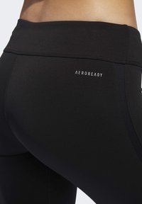 Lähikuva mustista urheilulegginsseistä, joissa on korkea vyötärö ja pieni "AEROREADY"-logo lonkalla.