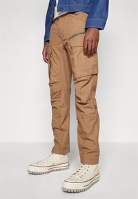 Kha ki cargobroek met meerdere zakken, een ritszak op de dij en een relaxte pasvorm. Draagt samen met crèmekleurige hoge sneakers.