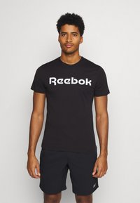 Svart bomull T-skjorte med hvit "Reebok" logo over brystet. Korte ermer, rund hals, kombinert med mørke shorts. En enkel, uformell design.