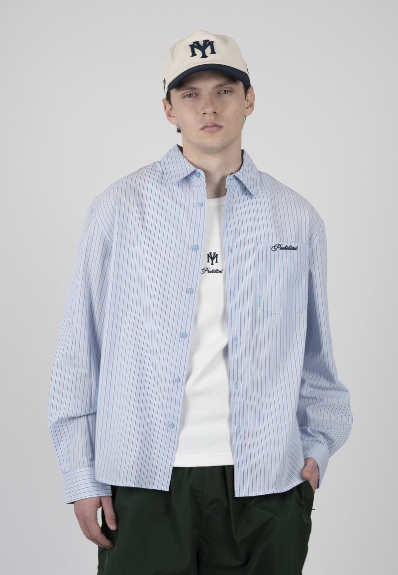 Jeune homme portant une casquette de baseball crème, une chemise rayée bleu et blanc à boutons par-dessus un t-shirt blanc avec logo, pantalon vert foncé, expression neutre.