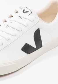 Valkoinen nahkainen lenkkari, jossa musta logo yksityiskohtana sivussa, litteät nauhat ja beige sisävuori. Kumipohja, jossa on kuvioitu pinta.