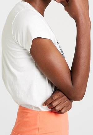 Witte T-shirt met korte mouwen en een relaxed fit, tentoongesteld naast een perzikkleurige hoge taille rok, met nadruk op de textuur van de stof en kleurcontrast.