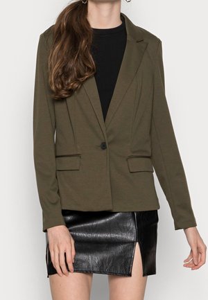 Kvinde iført en olivengrøn blazer over en sort top kombineret med et sort lædermininederdel med frontslids, stående mod en ensartet baggrund.
