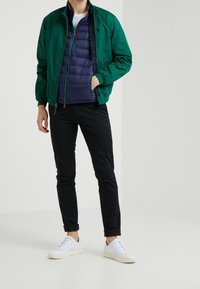 Zelená bomber bunda přes námořnickou plněnou vestu, černé kalhoty a bílé tenisky. Materiál je hladký s zipy a kapsami.