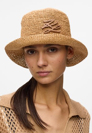 Jeune femme aux longs cheveux foncés portant un chapeau seau beige en osier avec un emblème décoratif marron clair et un haut beige assorti à mailles ouvertes.