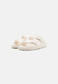 Hvite sandaler med to stropper som har justerbare spenner, teksturert såle og robust såle, designet for komfort og hverdagsbruk.