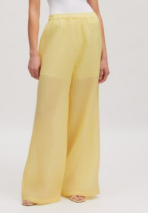 Frau trägt eine weit geschnittene, transparente, gelbe Hose mit elastischem Bund, kombiniert mit einem weißen Oberteil und offenen Sandalen vor einfachem Hintergrund.