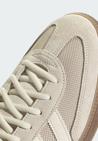Beige mockasinsneaker med meshpaneler, platta snören och texturerad gummisula. Inkluderar ljus sömnad och subtila vinklade linjer.