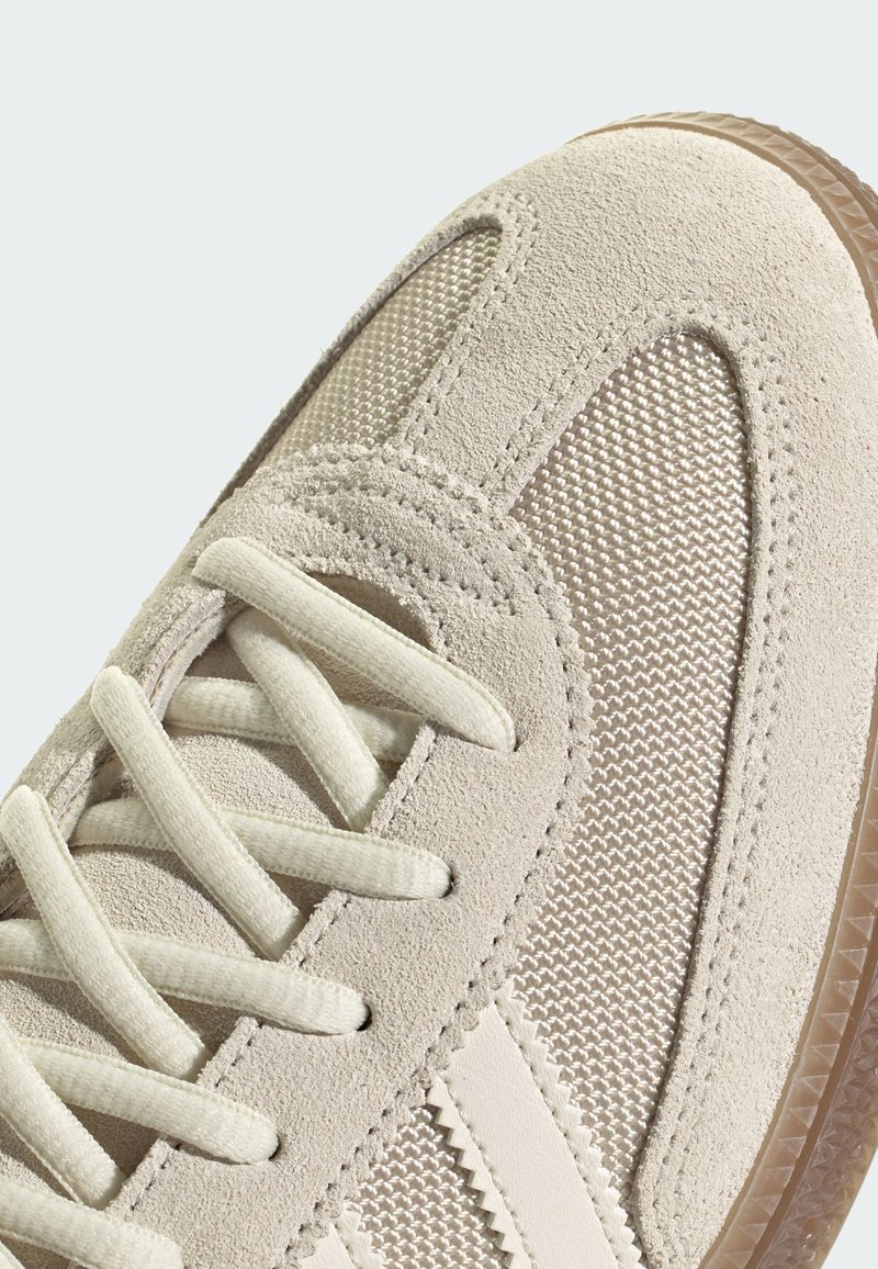Beige mockasinsneaker med meshpaneler, platta snören och texturerad gummisula. Inkluderar ljus sömnad och subtila vinklade linjer.