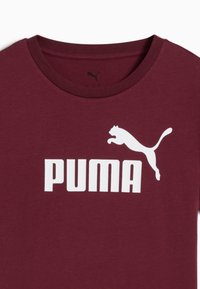 Burgundska pamučna majica s okruglim izrezom, koja ima veliki bijeli "PUMA" logo i grafiku skakačke mačke. Glatka tekstura.