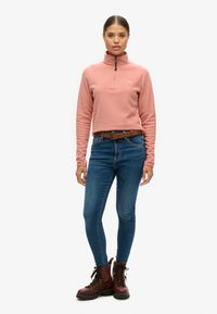 Pullover in pile rosa chiaro con zip a metà, jeans in denim blu e stivali marroni con lacci e suola in gomma. Branding minimale sul pullover.