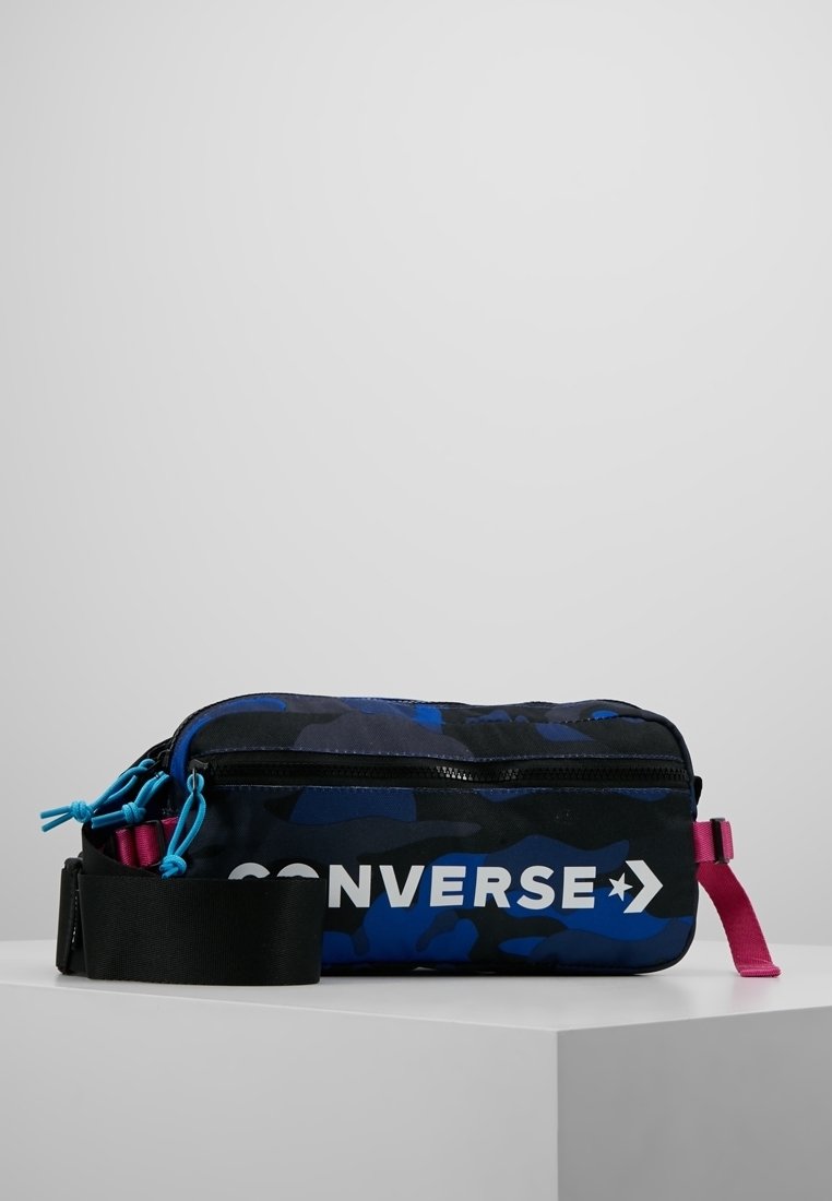 Sac bandoulière rectangulaire camouflage bleu et noir avec sangle noire, tirettes de fermeture éclair roses et bleues, et logo Converse blanc sur le devant.