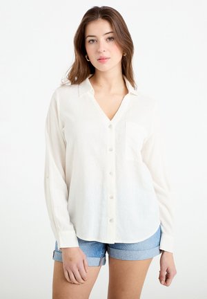 Joven con cabello castaño claro, vistiendo una camisa blanca abotonada y pantalones cortos de mezclilla arremangados, con un fondo blanco liso.