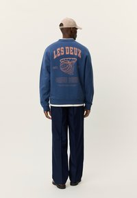 Μπλε φούτερ με την επιγραφή «LES DEUX» σε πορτοκαλί γράμματα στην πλάτη, με γραφικό θέμα μπάσκετ και με λευκό πουκάμισο από κάτω.