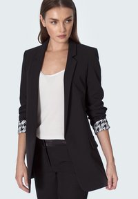 Kobieta ubrana w czarną marynarkę z podszewką w wzór houndstooth, białą bluzkę bez rękawów i czarne spodnie, patrzy w lewo na tle jednolitego tła.