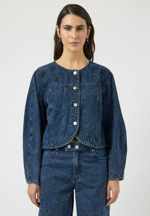 Femme portant une veste en denim bleu foncé et un pantalon assorti avec des broderies florales, debout devant un fond clair uni.