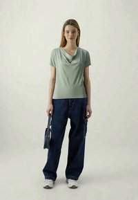 Top de manga curta verde-claro com decote drapeado, combinado com jeans soltos azul-escuro e ténis brancos. Segura uma pequena bolsa cinza.