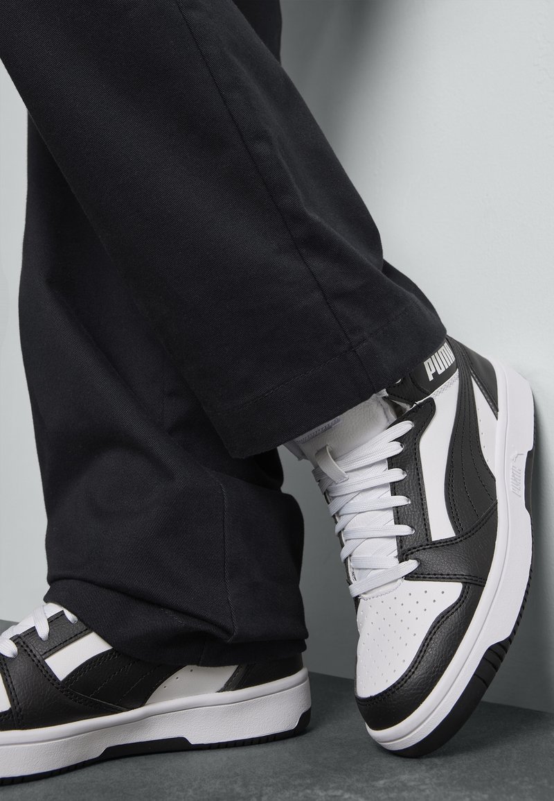 Personne portant des baskets Puma noires et blanches et un pantalon noir, debout avec un pied légèrement levé contre un mur gris.
