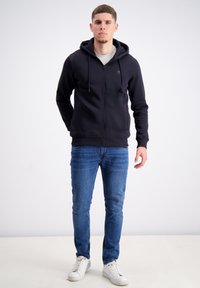 Marineblå zip-up hoodie med snoretræk, parret med lyseblå jeans og hvide sneakers. Stoffet ser blødt og glat ud, hvilket skaber et afslappet look.