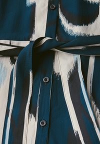 Camicia button-up con un motivo astratto in turchese, nero e crema. Realizzata in tessuto morbido con un dettaglio in vita annodato e bottoni scuri.