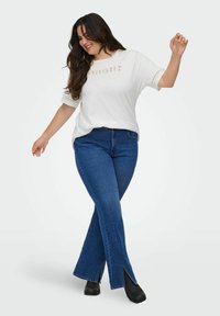 T-shirt bianco a maniche corte con dettaglio testuale "Amour", abbinato a jeans a campana blu scuro con fessure laterali e stivaletti neri.