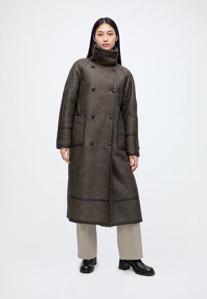 Un mannequin se tient devant un fond blanc, vêtu d’un long manteau marron foncé à col montant, d’un pantalon beige et de bottines noires.