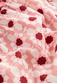 Textilie s geometrickým vzorem, který obsahuje světle růžové, tmavě růžové a burgundské tvary. Látka má hladkou, jemnou strukturu.