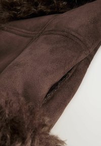 Material din suede maro, cu o textură moale. Are o buzunar lateral și accente pufoase maro pe margine, indicând căldură și confort.