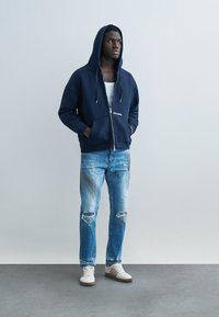 Muž s rukami v vreckách, oblečený v tmavomodrej mikine na zips, bielom tielku, roztrhaných svetlomodrých džínsach a bielych teniskách stojaci pri jednoduchej stene.