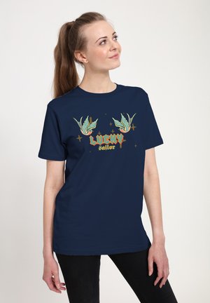 Ung kvinde med langt hår iført navyblå T-shirt med to lyseblå fugle og teksten "Lucky Sailor". Hun kigger opad med afslappede hænder.