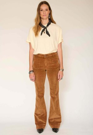 Haut à manches courtes crème associé à un pantalon en velours côtelé marron évasé. Écharpe noire et blanche autour du cou, bracelet épais au poignet.