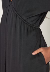 Schwarzes Kleid mit gerafftem Taillengebiet, kurzen Ärmeln und einer Tasche auf einer Seite. Der Stoff ist weich und hat eine glatte, fließende Textur.