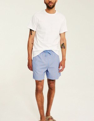 Mand iført en enkel hvid t-shirt, blå og røde stribede shorts, brune sandaler, med synlige tatoveringer på arme og ben, stående på en lys baggrund.