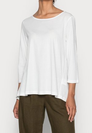 Vrouw draagt een losse witte top met lange mouwen en bootnek tegen een effen achtergrond, gecombineerd met donkerbruine broek.